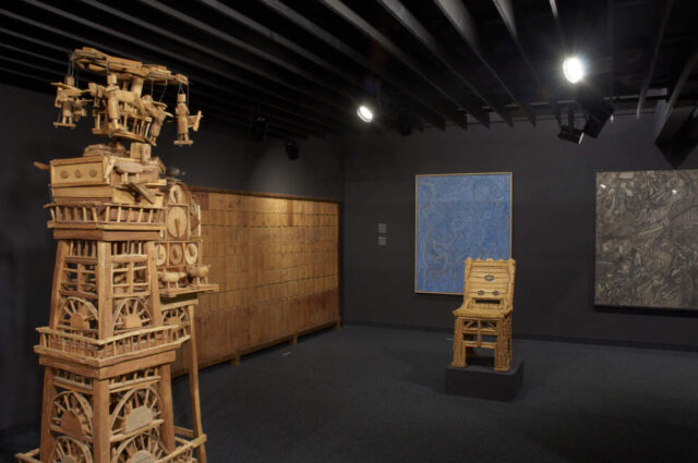 Blick in einen Ausstellungsraum. Im Vordergrund steht eine Turmskulptur aus Holz.