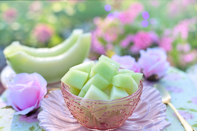 Hellgr&uuml;ne Honigmelonenw&uuml;rfel in einer rosafarbenen, gemusterten Glassch&uuml;ssel auf einem passenden rosafarbenen Glasteller. Im Hintergrund zwei Melonenspalten und rosa Bl&uuml;ten in einem unscharfen, hellen Aussensetting.