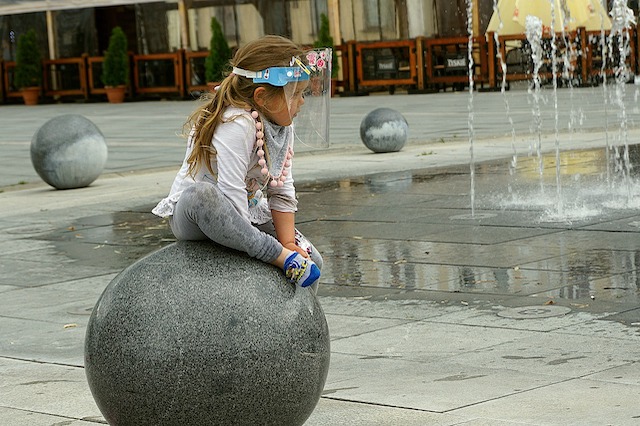 Ein kleines Mädchen sitzt auf einer grossen Steinkugel. Sie trägt ein Visier mit einem Gummiband.