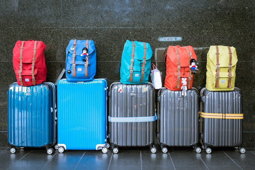Fünf Rollkoffer stehen nebeneinander. Auf jedem Rollkoffer steht ein Reiserucksack. Jeder Rucksack hat eine andere Farbe.