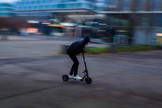 Eine Person in dunkler Kapuzenjacke und heller Schuhe f&auml;hrt in stark vorgebeugter Haltung auf einem schwarzen E-Scooter &uuml;ber einen weiten, asphaltierten Platz; die Bewegungsunsch&auml;rfe betont die hohe Geschwindigkeit.