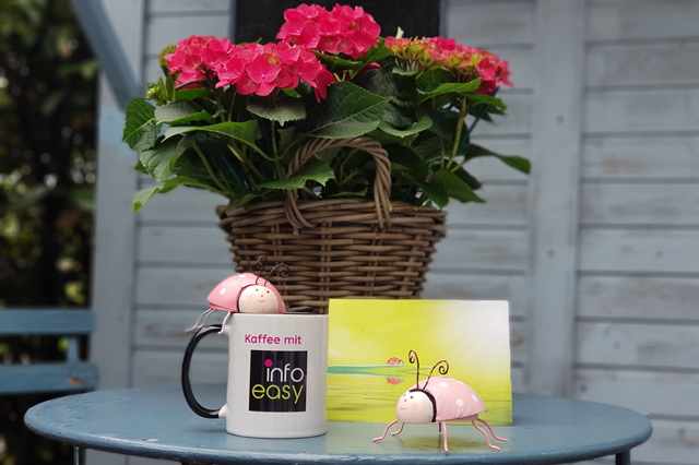 Auf einem Gartentischchen steht eine Tasse mit dem infoeasy Logo. Auf der Tasse sitzt ein rosaroter Marienkäfer aus Blech. Ein zweiter solcher Marienkäfer steht neben der Tasse.
