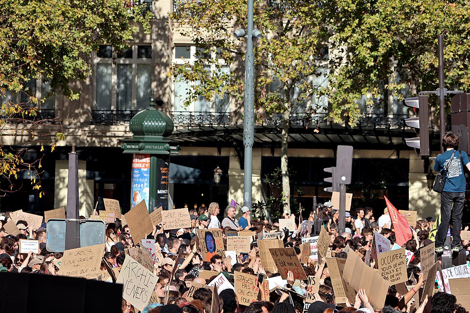 Eine grosse Menschenmenge bei einer Demonstration. Viele Personen halten Schilder aus brauner Pappe mit franz&ouml;sischen Texten hoch. Rechts steht eine Person erh&ouml;ht an einem schwarzen Ampelmast. Im Hintergrund stehen B&auml;ume, eine dunkelgr&uuml;ne Litfasss&auml;ule und ein helles Geb&auml;ude.