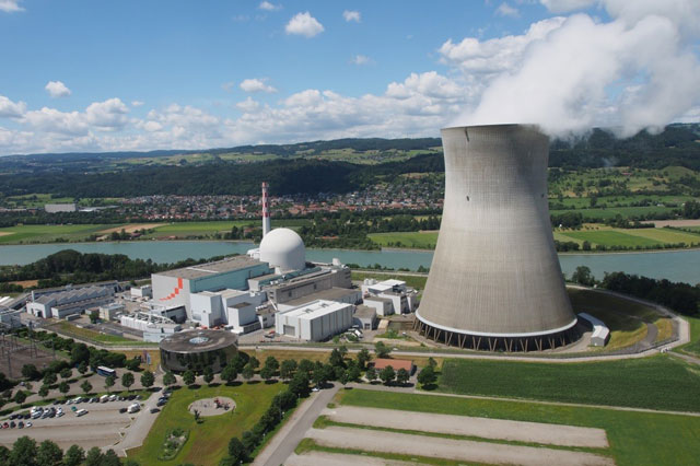 Eine Luftaufnahme des Kernkraftwerks Leibstadt: : ein gro&szlig;er hyperbolischer K&uuml;hlturm aus Beton mit aufsteigendem Wasserdampf, daneben das wei&szlig;e Reaktorgeb&auml;ude mit charakteristischer Kuppel. Die Anlage liegt an einem Fluss, umgeben von Feldern und einer bewaldeten H&uuml;gellandschaft.