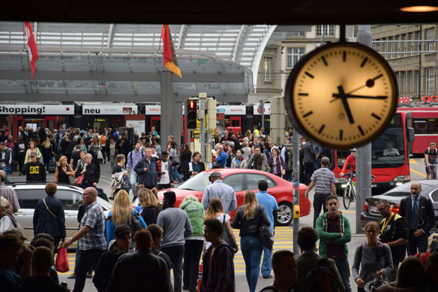 Belebter Bahnhofsvorplatz mit zahlreichen Fussg&auml;ngern, einem roten Auto und einem roten Bus; im Vordergrund rechts eine h&auml;ngende runde Bahnhofsuhr mit goldenem Zifferblatt; im Hintergrund ein Glasdach, Nationalflaggen sowie der Werbeschriftzug &laquo;shopping! neu&raquo;.