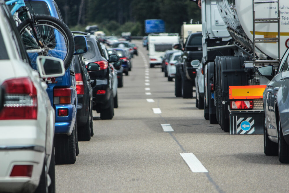 Dichter Stau auf einer mehrspurigen Autobahn: links eine Reihe von Pkw (darunter ein blauer SUV mit Fahrradtr&auml;ger auf dem Dach), rechts ein schwerer Lkw-Sattelzug mit orangerotem R&uuml;ckreflektor und einem Hinweisschild mit blauem Pfeil. Zwischen den Fahrzeugkolonnen eine gestrichelte Mittellinie; im Hintergrund ein blaues Autobahnschild und Baumreihen.