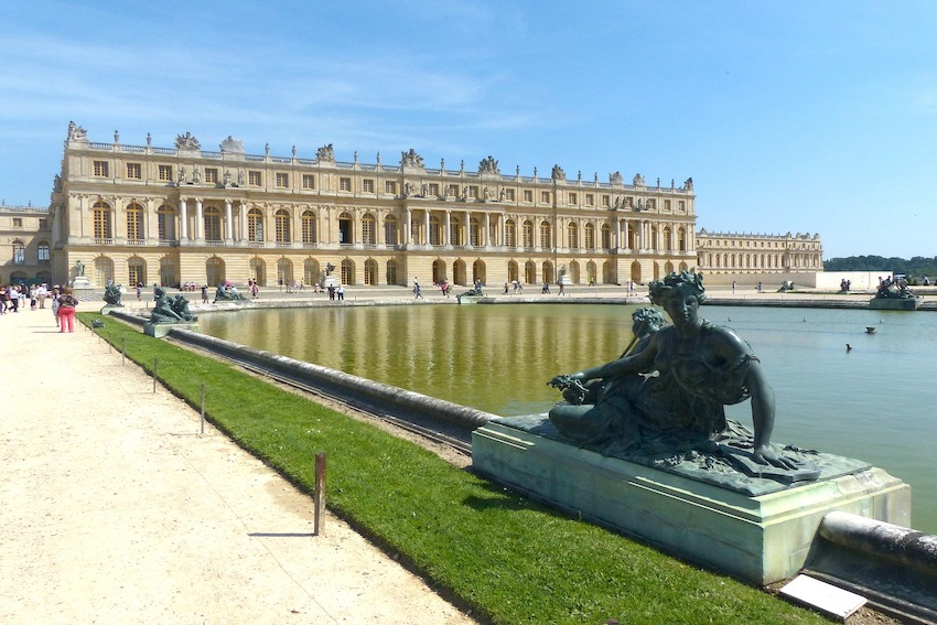Im Hintergrund ist eine grosse Fassade von Schloss Versailles. Im Vordergrund gibt es ein grosses Wasserbecken. In der Mauer des Wasserbeckens sind in Abständen Statuen eingearbeitet. Am linken Bildrand ist ein Kiesweg, auf dem Touristen laufen und stehen.