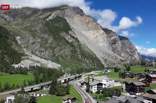 Luftaufnahme des Dorfes Randa im Oberwallis mit dem Logo-Schriftzug SRF news oben links. Im Hintergrund ein massiver Berghang mit grossfl&auml;chiger Felssturzfl&auml;che aus hellem Gestein; darunter gr&uuml;ne Wiesenh&auml;nge. Im Dorf verlaufen Strasse und Bahntrasse mit einem roten Zug parallel durch die Ortschaft mit traditionellen Holzh&auml;usern. ALT-Text erstellt mit inkludocs.