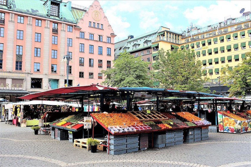 Eine Szene auf einem Markt in Stockholm. Vorne ist ein Gemüse- und Früchtestand. Dahinter ist ein Stand mit Kleidern.