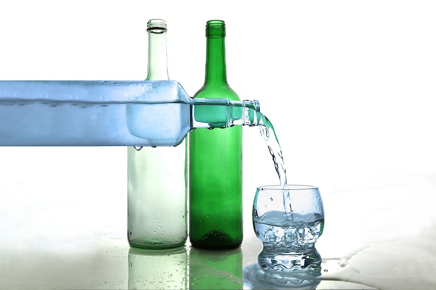 Im Hintergrund stehen eine weisse und eine grüne Glasflasche. Beide Flaschen sind leer. Im Vordergrund wird aus einer weissen Glasflasche Wasser in ein Glas gefüllt.
